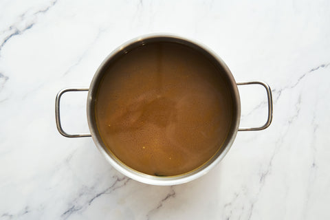 Overhead photograph of a stainless steel pot containing rich brown broth resting on a white marble surface with subtle grey veining