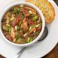 A photograph of a hearty chicken and sausage gumbo with green onions and celery in a dark roux served in a white bowl alongside a slice of golden garlic toast on a rustic wooden table