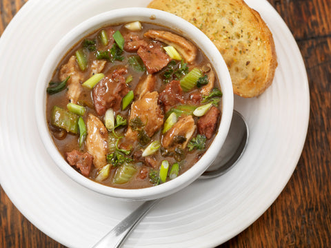 A photograph of a hearty chicken and sausage gumbo with green onions and celery in a dark roux served in a white bowl alongside a slice of golden garlic toast on a rustic wooden table
