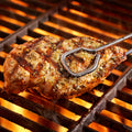Photography of a marinated chicken breast held by metal tongs being grilled over glowing orange flames, showing distinct char marks, green herbs, and rising smoke