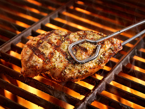 Photography of a marinated chicken breast held by metal tongs being grilled over glowing orange flames, showing distinct char marks, green herbs, and rising smoke