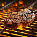 A mouth-watering, perfectly seared steak with prominent grill marks and cracked pepper seasoning is being turned by metal tongs over a hot barbecue grill with vibrant orange flames