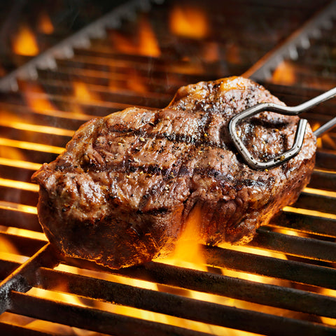 A mouth-watering, perfectly seared steak with prominent grill marks and cracked pepper seasoning is being turned by metal tongs over a hot barbecue grill with vibrant orange flames