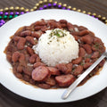 A classic Cajun red beans and rice dish with sliced Andouille sausage and herb garnish on a white plate, set on a rustic wooden table alongside vibrant purple, green, and gold Mardi Gras beads