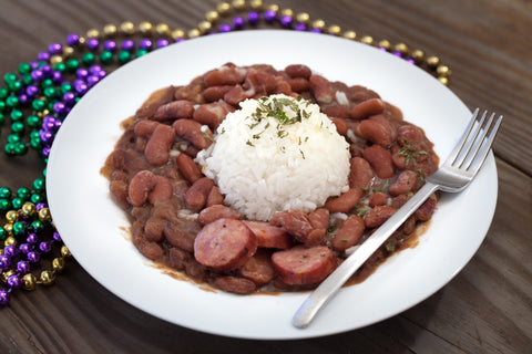A classic Cajun red beans and rice dish with sliced Andouille sausage and herb garnish on a white plate, set on a rustic wooden table alongside vibrant purple, green, and gold Mardi Gras beads