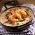 Photography of plump, seasoned shrimp and creamy grits in a rustic skillet, topped with crispy bacon and fresh green parsley, resting on a purple and white patterned napkin on a distressed wooden surface