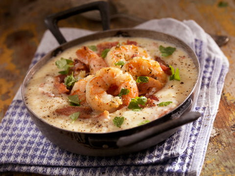 Photography of plump, seasoned shrimp and creamy grits in a rustic skillet, topped with crispy bacon and fresh green parsley, resting on a purple and white patterned napkin on a distressed wooden surface