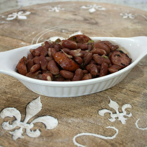 Photograph of a hearty dish of red beans and sliced charred sausage in a white ceramic bowl on a wooden table with white fleur-de-lis patterns evoking New Orleans cuisine