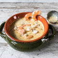 Photograph of creamy seafood bisque with plump pink shrimp and white crab meat served in a rustic green and terra cotta ceramic bowl on a distressed white wooden surface, a brass ladle resting beside