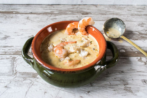 Photograph of creamy seafood bisque with plump pink shrimp and white crab meat served in a rustic green and terra cotta ceramic bowl on a distressed white wooden surface, a brass ladle resting beside