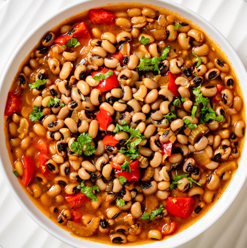 Top-down view of a flavorful black-eyed pea stew in a white bowl, featuring tender peas, bright red bell peppers, and fresh green herbs in a rich orange broth
