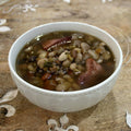 Photograph of a hearty white bowl of mixed bean and smoked ham soup garnished with green herbs on a rustic wooden surface with faint white decorative flourishes