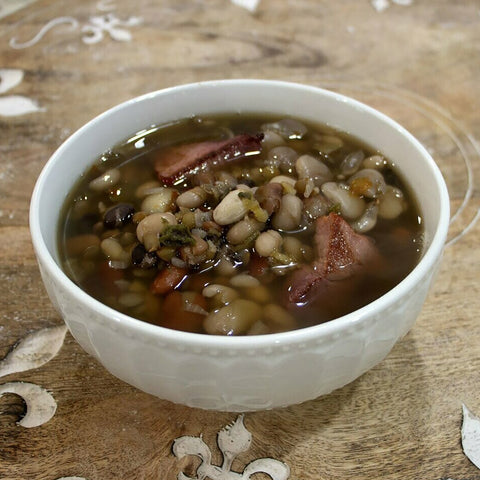 Photograph of a hearty white bowl of mixed bean and smoked ham soup garnished with green herbs on a rustic wooden surface with faint white decorative flourishes